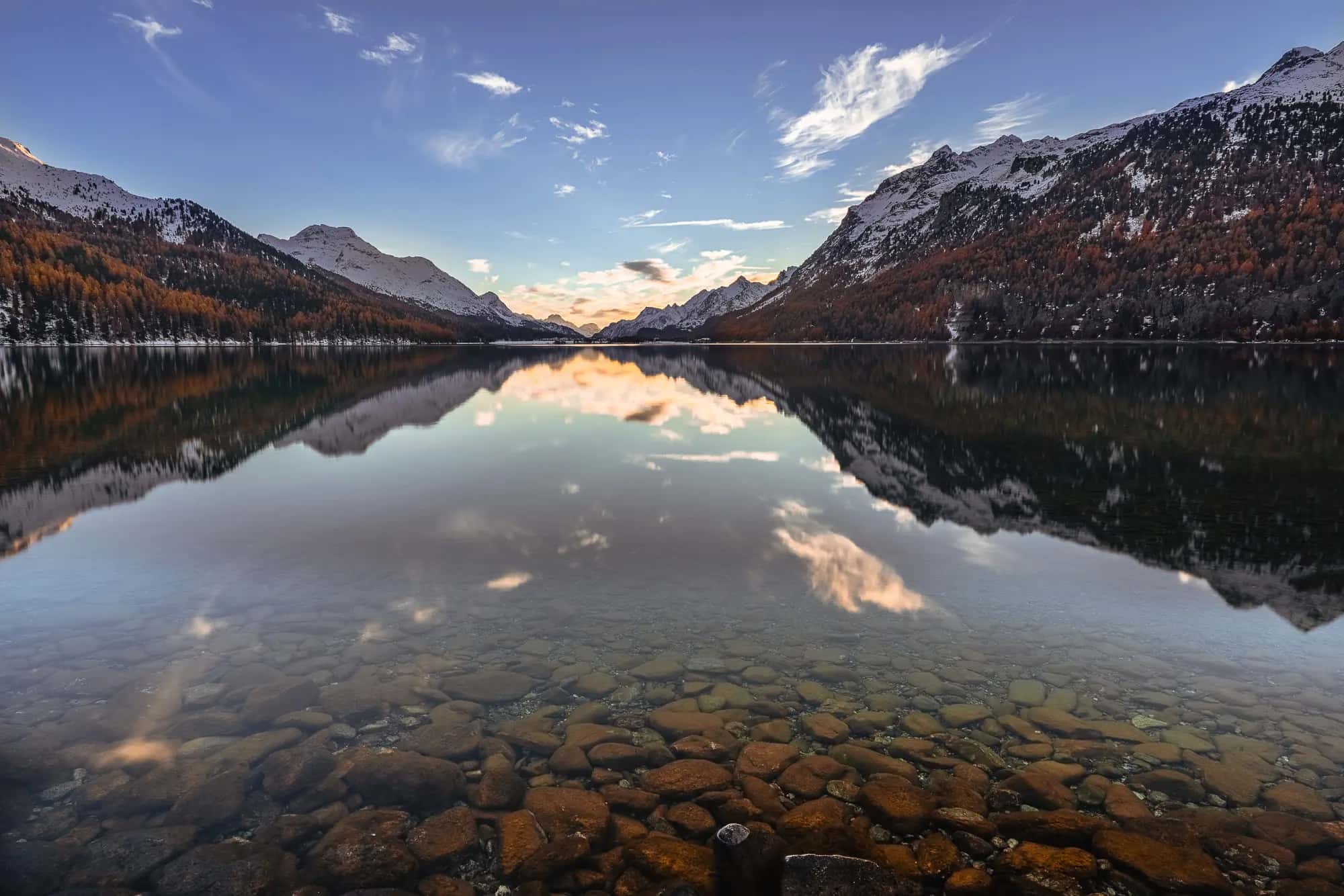 Ein interessanter Vordergrund leitet in die Spiegelung des Silvaplanasee über, die Berghänge sind mit Lärchen dekoriert und das gesamte Motiv läuft auf die Lichtstimmung am Schnittpunkt des Tals zu